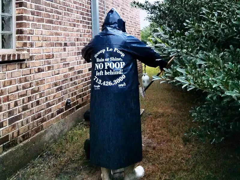 Scoop le Poop waste specialist working in the rain in a branded, dark blue rain slicker that says "Rain or Shine, no poop left behind" on the back.